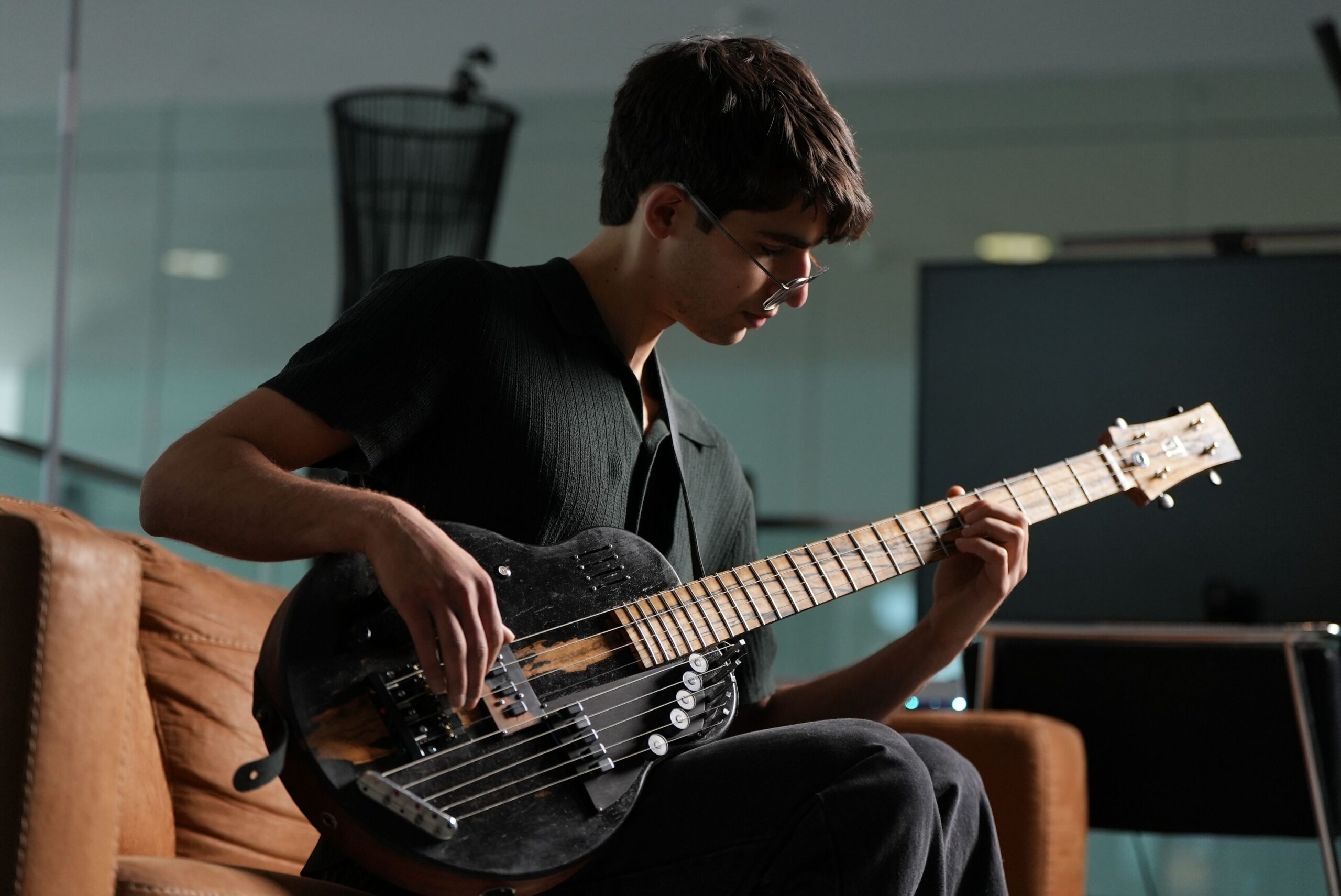 A person with short dark hair and glasses sits on a brown couch, playing a black electric bass guitar. The background is blurred, showing some modern office furniture and glass walls.