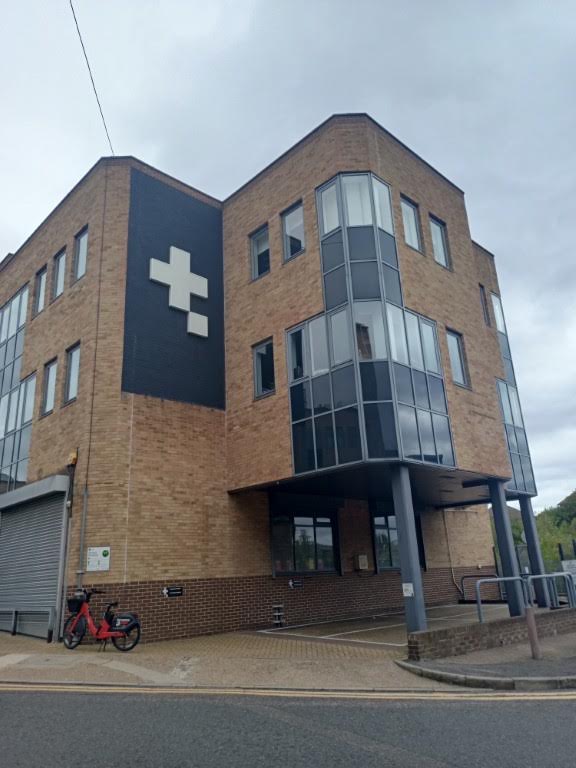 A brick building with a white cross logo on the side and a prominent glass-paneled corner, with a bicycle parked near the entrance.