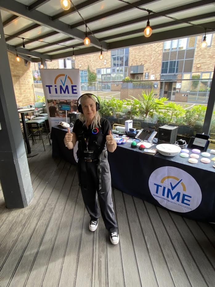A woman wearing a black headset and shirt stands at a booth for Technology in Music Education (TIME), giving a thumbs up behind a table covered with electronic equipment.