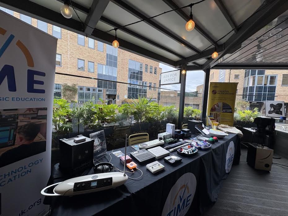 An outdoor table draped with a black cloth displaying various music technology devices, flanked by two informational standing banners.