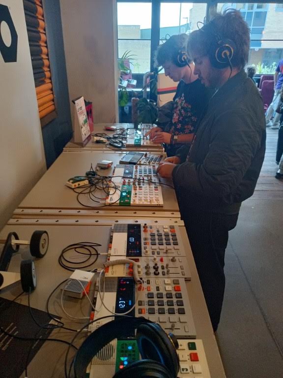 Two men standing at a long table, each wearing headphones and operating digital music samplers.