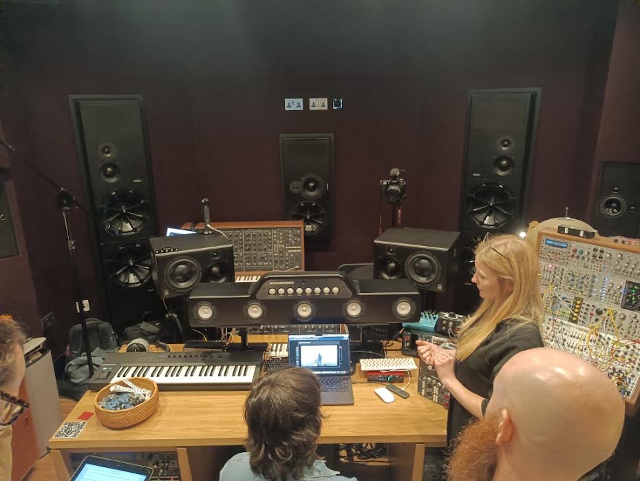 Three people in a music recording studio working at a desk filled with synthesizers, audio monitors, and computer equipment.