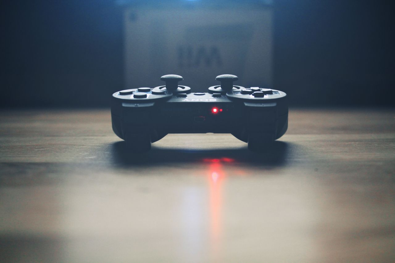 A black video game controller sits on a wooden surface with a small red light glowing on it, casting a faint red reflection. The background is blurred and dimly lit.