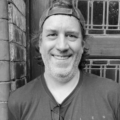 A smiling man wearing a baseball cap and a graphic t-shirt with eyeglasses tucked into the neckline.