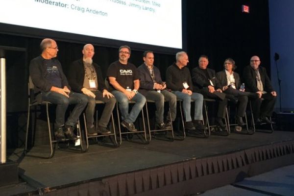 Eight men sit on high chairs on a stage during a panel discussion, with a large screen displaying names and a moderator behind them. They appear to be engaged in conversation at a professional event.