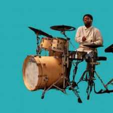 A drummer playing on a beige drum set in front of a solid turquoise background, wearing a white long-sleeve shirt, with a joyful expression while striking the drums.